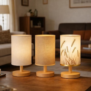 May include: Three table lamps with cylindrical shades and wooden bases. One shade features a wheat stalk design. The lamps are on a dark wooden table. The lamps are in a living room.