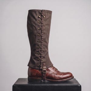 Puede incluir: Una polaina de tela marrón hasta la rodilla con un diseño de cordones vertical con ojales y ganchos en tono latón, asegurada en el tobillo con una correa de cuero marrón a juego y una hebilla en tono latón, sobre una bota de cuero marrón.