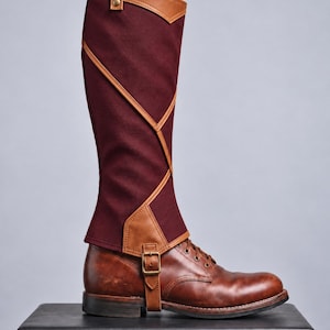 May include: A knee-high burgundy fabric gaiter with tan faux leather trim and geometric paneling, featuring a top strap with a metal snap and a lower adjustable buckle strap, worn over a brown leather lace-up boot with a low heel.