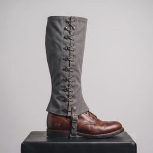 May include: A knee-high grey canvas gaiter featuring a vertical lace-up design with metal eyelets and hooks, secured by a brown leather strap and buckle at the ankle, worn over a brown leather lace-up boot with a low heel and a rounded toe.