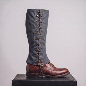 May include: A knee-high dark gray canvas gaiter featuring a vertical side lacing system with brown leather laces and brass eyelets, secured over a brown leather lace-up boot with a single adjustable leather strap and brass buckle at the ankle.