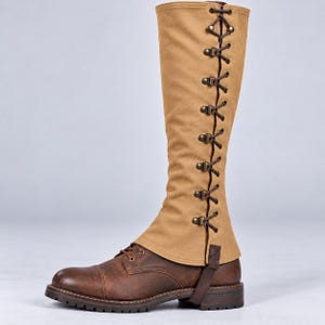 May include: A knee-high tan fabric gaiter featuring a vertical row of dark brown laces threaded through metallic eyelets, worn over a brown leather boot with a rounded toe, a low heel, and a thick, textured rubber sole for traction.