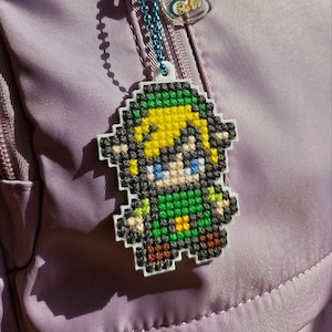 Può includere: Un ciondolo pixelato raffigurante un personaggio con capelli biondi, una tunica verde e occhi blu. Il ciondolo è attaccato a una catena blu e a un'etichetta di plastica bianca. Lo sfondo è un tessuto viola.