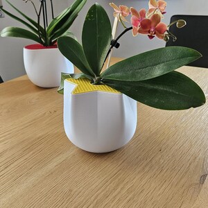 Può includere: Due vasi bianchi per orchidee con fiori rosa e arancioni. I vasi hanno un design geometrico e sono su un tavolo di legno chiaro. Le orchidee aggiungono un tocco di bellezza naturale all'ambiente interno.