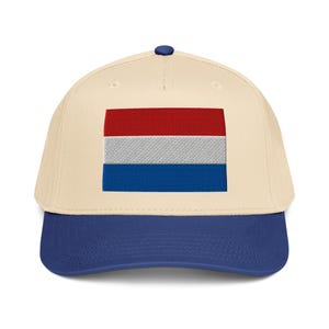 Peut inclure: Casquette de baseball avec une couronne crème et une visière bleu marine. Le devant de la casquette présente un drapeau brodé des Pays-Bas, avec des rayures horizontales rouges, blanches et bleues.