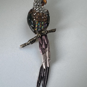Könnte beinhalten: Eine Papageienbrosche auf einem strukturierten, goldfarbenen Metallzweig. Der Vogel ist mit klaren, bernsteinfarbenen und irisierenden Strasssteinen an Kopf, Brust und Flügeln besetzt und hat lange, geschichtete Schwanzfedern in Violett und Silber.
