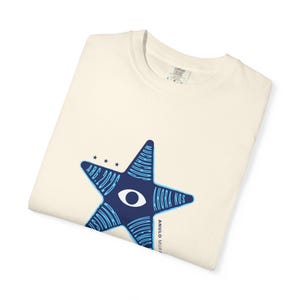 May include: Cream-colored t-shirt with a large blue star design. The star features an eye and text that reads "ARG.2026" and "AHORA NOS MUCHAADOS". The shirt is folded neatly.