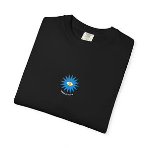 以下が含まれることがあります： 黒いTシャツ。青と黄色のスタイリッシュな目のグラフィックと「ANULO MUFA」の文字が特徴です。Tシャツは折りたたまれており、襟にはタグが付いています。