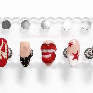 Könnte beinhalten: Eine transparente Acryl-Nagelpräsentation mit fünf einzigartigen Nageldesigns. Die Nägel zeigen ein rotes Design mit goldenen Akzenten, einen nudefarbenen Nagel mit schwarzen Nieten, ein Lippendesign, einen Nagel mit roten Sternen und einen Zebrastreifen-Nagel mit goldenen Akzenten. Der Text "@SOPHFREAKYNAILS" ist sichtbar.