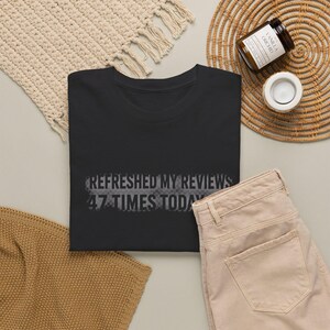 May include: A black t-shirt featuring the text "REFRESHED MY REVIEWS 47 TIMES TODAY" printed in a dark gray, distressed, pixelated font across the center of the chest. The shirt has a crew neckline and short sleeves, presented in a flat layout.