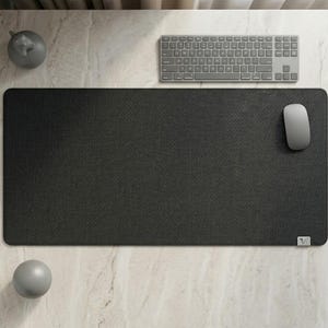 Puede incluir: Una alfombrilla de escritorio negra rectangular grande con una superficie texturizada. Un teclado y un ratón inalámbricos grises están colocados sobre la alfombrilla. Un pequeño objeto decorativo gris y una esfera gris también están sobre la superficie de mármol.