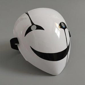 May include: A glossy white mask featuring a stylised black graphic design. The mask displays a wide, curved black smile, a single black teardrop-like line extending from the right eye, and a small blue accent within the right eye socket.