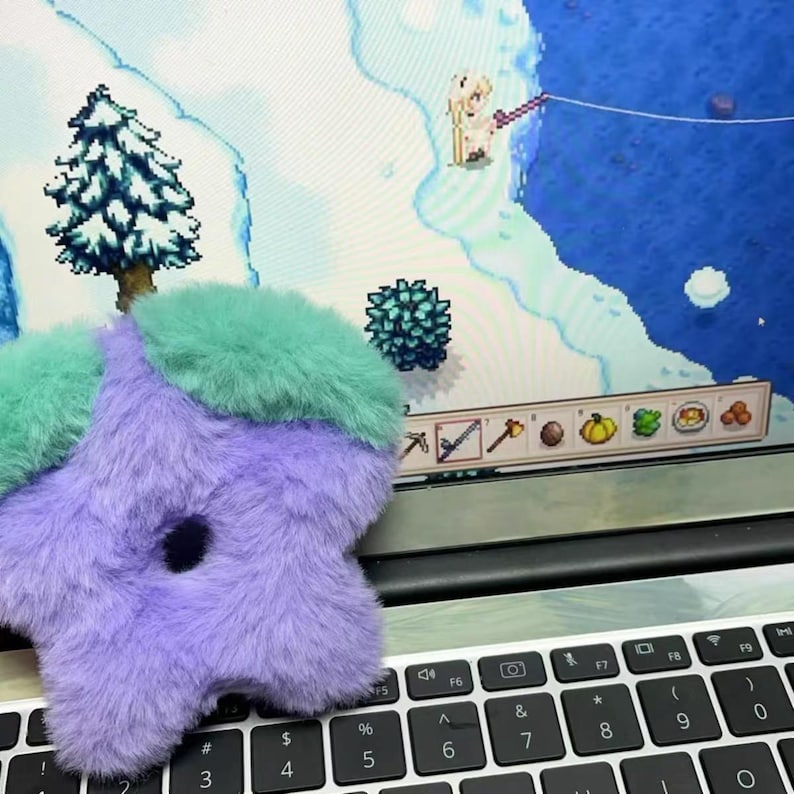 K&ouml;nnte beinhalten: Ein flauschiges, sternf&ouml;rmiges Accessoire in Lila und T&uuml;rkis, das auf einer Laptop-Tastatur liegt. Die Tastatur ist schwarz, und der Bildschirm zeigt eine pixelige Videospielszene mit einer verschneiten Landschaft.