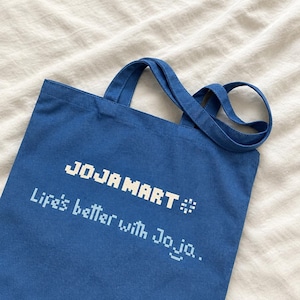 Könnte beinhalten: Eine blaue Tragetasche aus Segeltuch mit dem weißen, verpixelten Text JOJAMART und dem Slogan Life's better with Joja. Die Tasche hat zwei passende blaue Stoffgriffe an der Oberseite, was ein einfaches und funktionales Design zum Tragen von Gegenständen bietet.