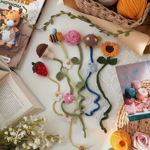 Op de afbeelding: Een verzameling handgemaakte gehaakte bladwijzers in verschillende bloemen- en natuurgeïnspireerde ontwerpen. De bladwijzers omvatten een aardbei, paddenstoel, bij, roos, zonnebloem en andere bloemmotieven. De bladwijzers zijn gerangschikt op een wit oppervlak met boeken en garen op de achtergrond.