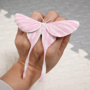 May include: A handmade crochet butterfly in pink and white. The butterfly has large, detailed wings with alternating stripes of pink and white. Long, delicate streamers extend from the body. The butterfly is held in a hand.