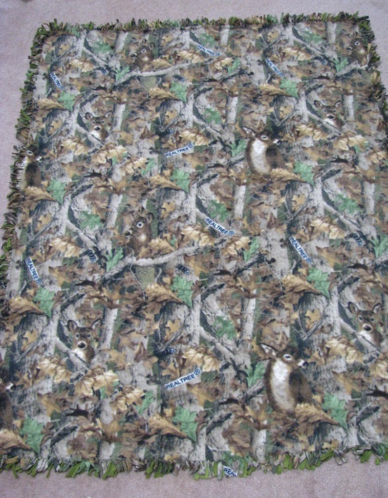 May include: A camouflage fleece blanket with a repeating pattern of deer and the words "RealTree" throughout. The blanket has a fringed edge.