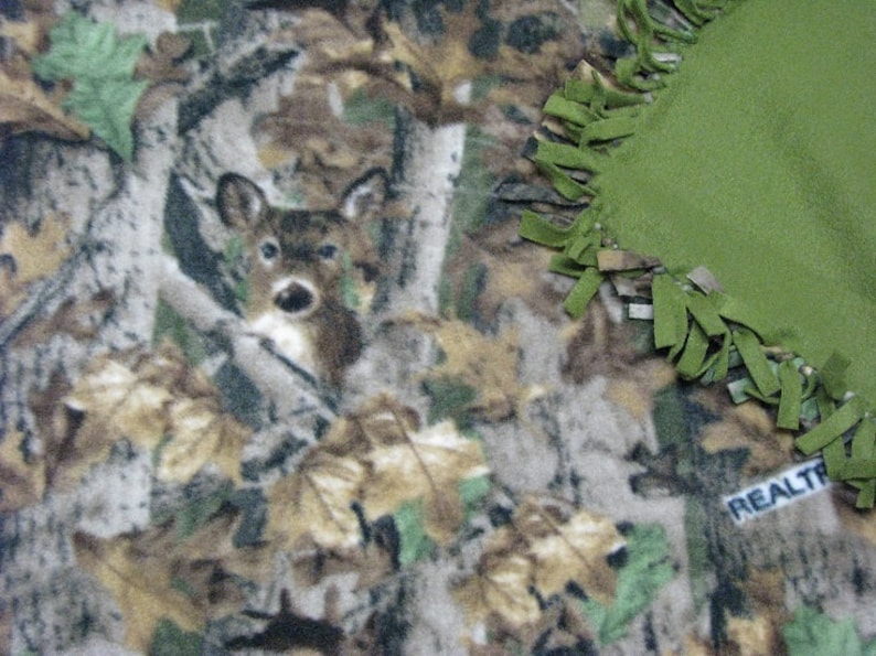 May include: A green and brown camouflage fleece blanket with a deer print and a green fringe border. The blanket has the word "REALTREE" printed on it.