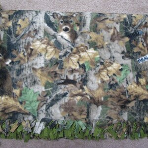 May include: A green and brown camouflage fleece blanket with a deer print and the text "REALTREE&reg;" in the upper right corner. The blanket has a fringe border.