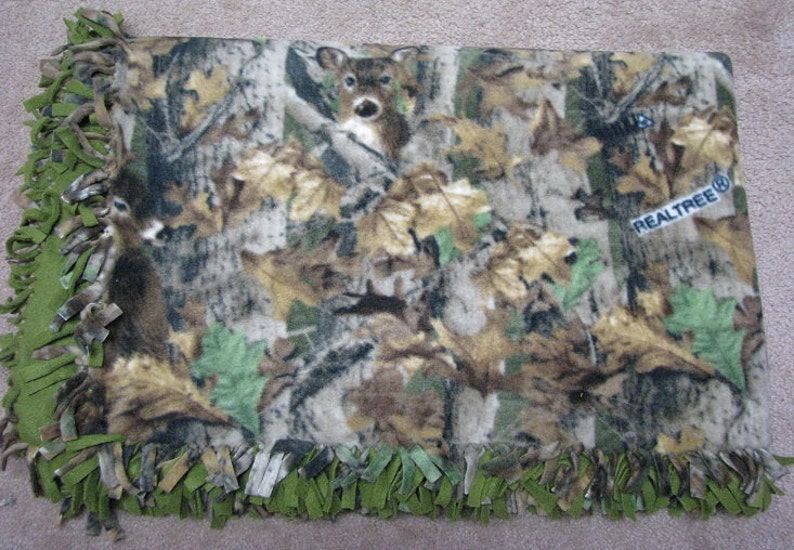 May include: A green and brown camouflage fleece blanket with a deer print and the text "REALTREE&reg;" in the upper right corner. The blanket has a fringe border.