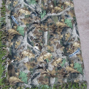 May include: A green, brown, and tan fleece blanket with a camouflage pattern featuring deer and the words "RealTree" printed on it. The blanket has a green fringe border.