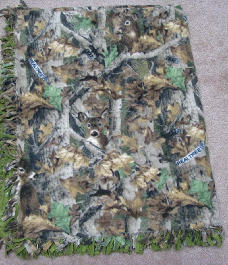 May include: A green, brown, and tan fleece blanket with a camouflage pattern featuring deer and the words "RealTree" printed on it. The blanket has a green fringe border.