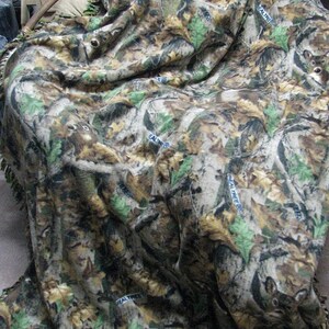 May include: A person is wrapped in a camouflage fleece blanket with a fringe border. The blanket has a brown, green, and tan color scheme with a repeating pattern of leaves and trees. The blanket has the words "Real Tree" printed on it.