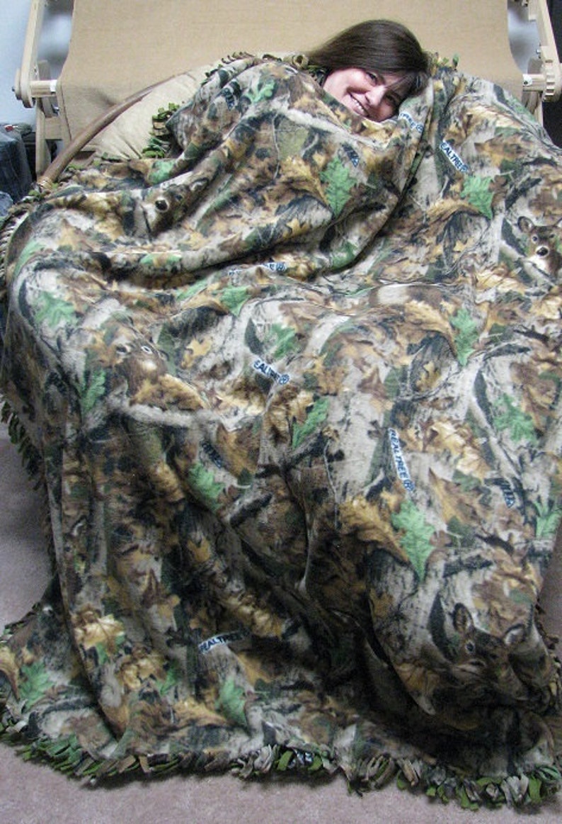 May include: A person is wrapped in a camouflage fleece blanket with a fringe border. The blanket has a brown, green, and tan color scheme with a repeating pattern of leaves and trees. The blanket has the words "Real Tree" printed on it.