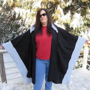 May include: A black and light blue fleece poncho with a hood. The poncho has a contrasting trim around the edges. The model is wearing the poncho over a red shirt and blue jeans.