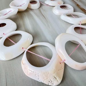 May include: A necklace featuring large, oval-shaped, off-white shell beads strung on a pink cord. The beads have a smooth, glossy finish with some showing natural textures and subtle peach-colored hues. The necklace is displayed on a light-colored surface.