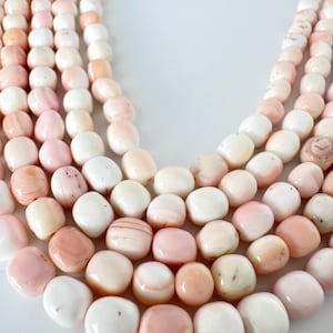 May include: A multi-strand necklace composed of smooth, rounded beads in varying shades of pale pink, cream, and white. The beads exhibit natural color variations and subtle textures, arranged in several cascading rows that create a layered look.