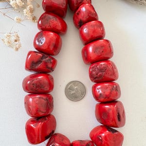 May include: A strand of large, irregular, bright red beads with natural surface textures and indentations. A silver-colored United States quarter coin is placed in the center of the strand for scale, providing a clear size reference for the beads.