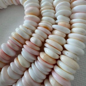 May include: A collection of smooth, disc-shaped beads arranged in multiple vertical strands. The beads are primarily white with subtle variations of soft pink and peach hues, featuring a polished, glossy finish and a uniform, rounded profile.