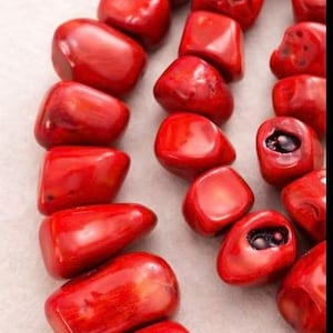 May include: Close-up of a strand of polished, vibrant red coral beads. The beads are irregularly shaped, with a glossy finish, and appear to be strung together. The background is a light, textured surface.