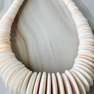 May include: A cream-colored necklace composed of numerous flat, rounded shell-like beads. The beads are strung together, forming a circular shape. The necklace has a natural, organic aesthetic, with a subtle pinkish hue visible on some beads.