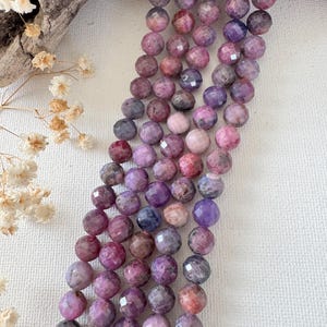 May include: Three strands of faceted round beads in shades of purple, magenta, and deep pink. The beads have a polished, multi-faceted surface that reflects light, creating a textured appearance across the strings of natural gemstone material.