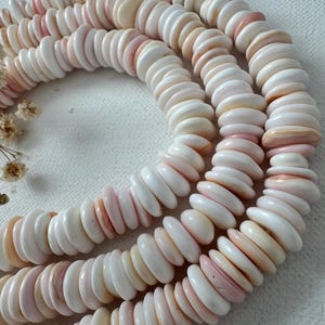 May include: A long strand of circular, flat shell beads arranged in a coiled pattern. The beads feature a smooth, polished texture with a color palette ranging from creamy white to soft, pale pink, showcasing natural variations in tone and shape.