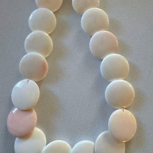 May include: A necklace composed of smooth, circular, flat beads in shades of off-white and pale pink. The beads are arranged in a single strand, featuring a consistent round shape and a polished finish, creating a uniform and simple jewelry piece.