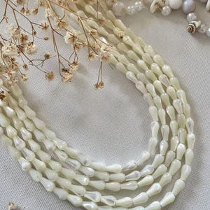 May include: A multi-strand necklace featuring off-white, teardrop-shaped beads with a pearlescent finish. The necklace is arranged in several curved rows, accompanied by dried baby's breath flowers and a cluster of small white shell beads.