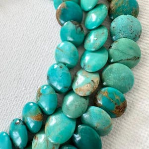May include: A strand of polished turquoise beads featuring a vibrant teal-blue color with natural brown matrix veining. The beads are oval-shaped and vary slightly in size and texture, creating a continuous, organic pattern of smooth, rounded stones.