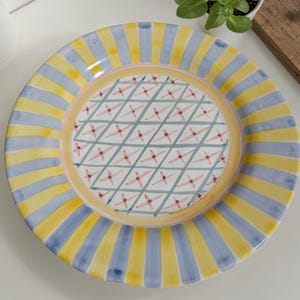 May include: A decorative ceramic plate with a colorful design. The plate features alternating yellow and blue vertical stripes around the rim. The center has a geometric pattern of intersecting lines and small red star-like shapes. A pink and yellow border surrounds the center design.