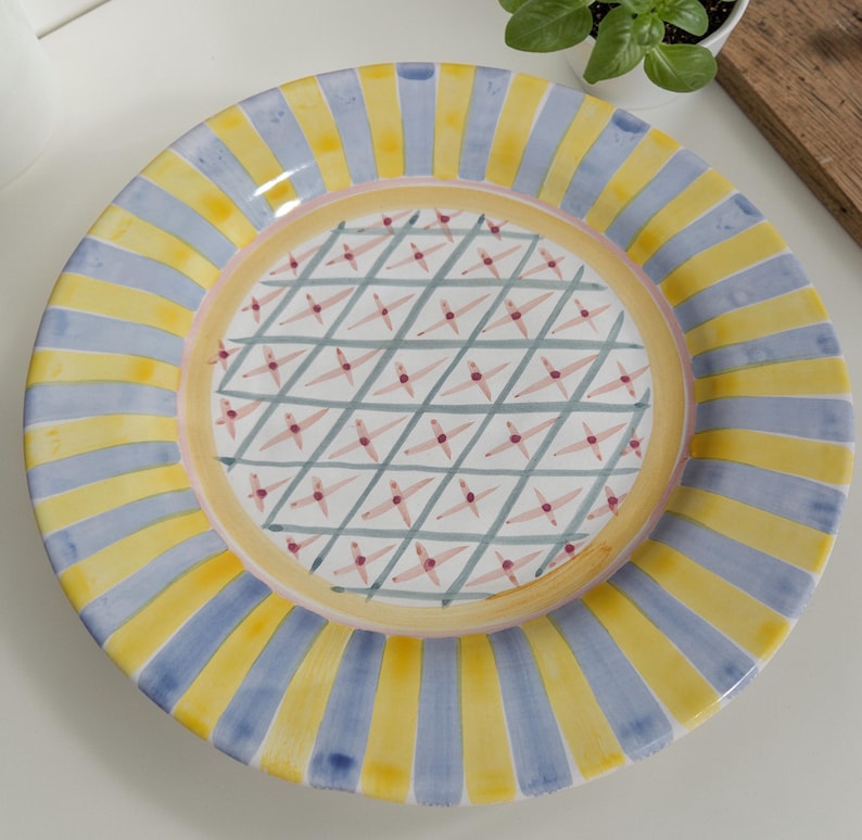 May include: A decorative ceramic plate with a colorful design. The plate features alternating yellow and blue vertical stripes around the rim. The center has a geometric pattern of intersecting lines and small red star-like shapes. A pink and yellow border surrounds the center design.