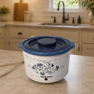 May include: A white Rival Crock-Ette slow cooker with a blue lid and a blue floral pattern on the exterior. The appliance features a round shape, a small power indicator light near the base, and an attached beige electrical power cord.