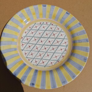 May include: A round ceramic plate with a decorative design. The rim features alternating yellow and light blue vertical stripes. The center has a pattern of intersecting lines with small red star-like shapes.