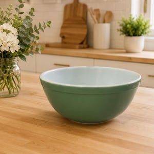 Puede incluir: Un cuenco verde y azul claro sobre una encimera de madera. El cuenco está vacío y tiene forma redondeada. Un tarro de cristal con flores blancas y vegetación está a la izquierda del cuenco. Una pequeña planta en maceta y utensilios de madera están al fondo.
