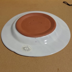 May include: A white ceramic plate with a wide rim and a terracotta-colored base. The plate has a slightly curved edge and a sticker on the bottom. The plate is likely for serving food.