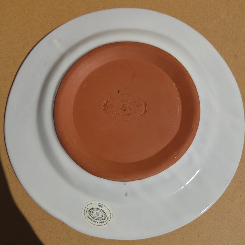May include: A white ceramic plate with a wide rim, viewed from the underside. The center is a reddish-brown color, with a stamped design. A small sticker is visible on the rim.