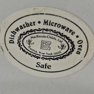 May include: Close-up of a white and oval sticker with black text. The sticker reads "Dishwasher &bull; Microwave &bull; Oven Safe" and includes the text "MacKenzie-Childs, Ltd. Aurora, New York 13026".