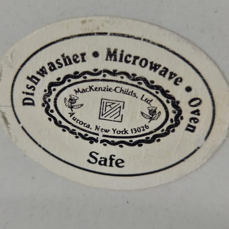 May include: Close-up of a white and oval sticker with black text. The sticker reads "Dishwasher &bull; Microwave &bull; Oven Safe" and includes the text "MacKenzie-Childs, Ltd. Aurora, New York 13026".