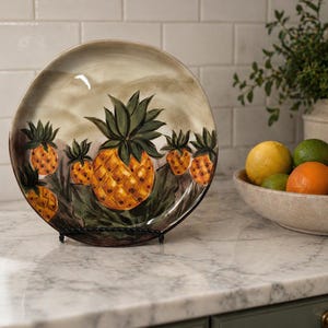 May include: A decorative plate featuring a hand-painted design of pineapples. The plate has a beige background with green and brown accents. The plate is displayed on a black metal stand, with a bowl of citrus fruits in the background.